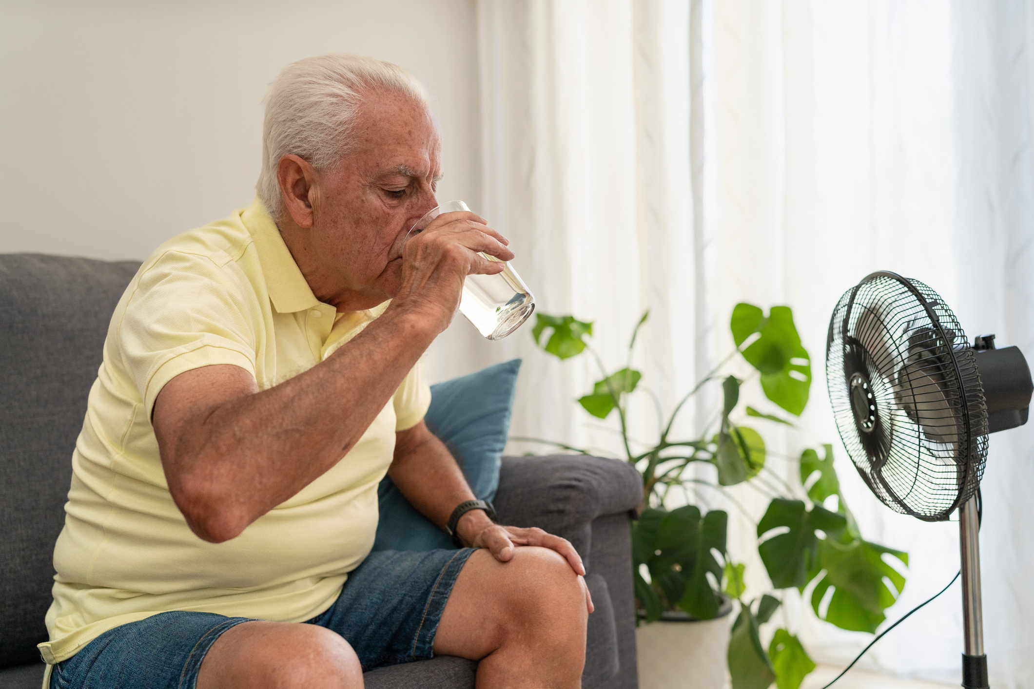 An older adult sitting in front of a fan and drinking water.
