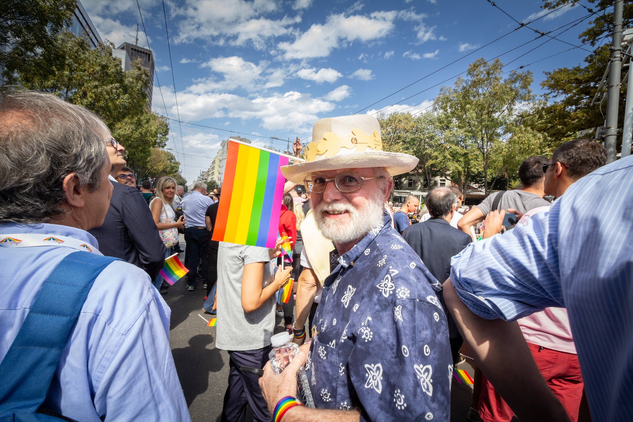 A man wearing a hat holds a rainbow flag, surrounded by a crowd celebrating pride