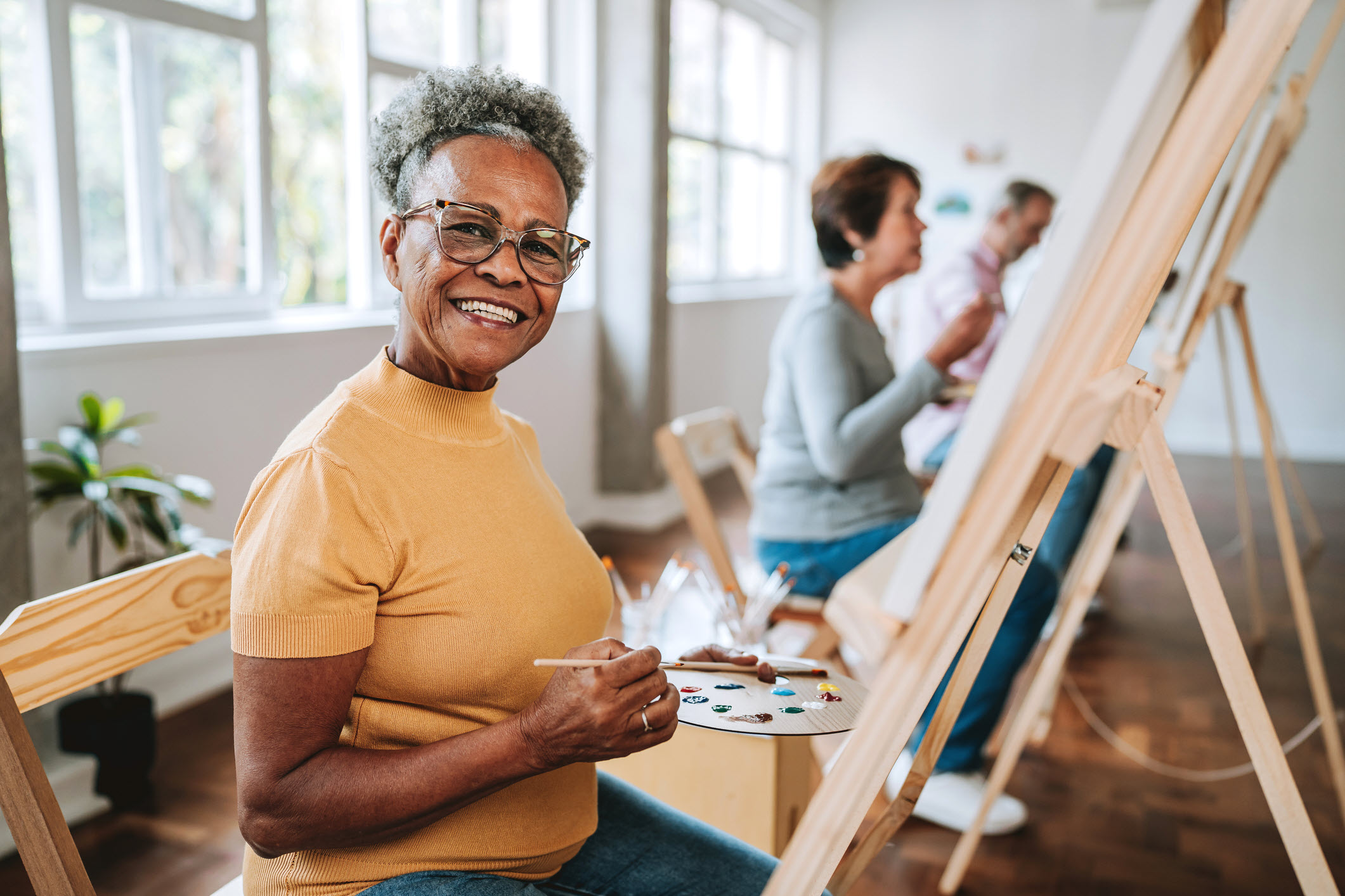Senior woman engaged in painting alongside peers in a lively, communal art environment.