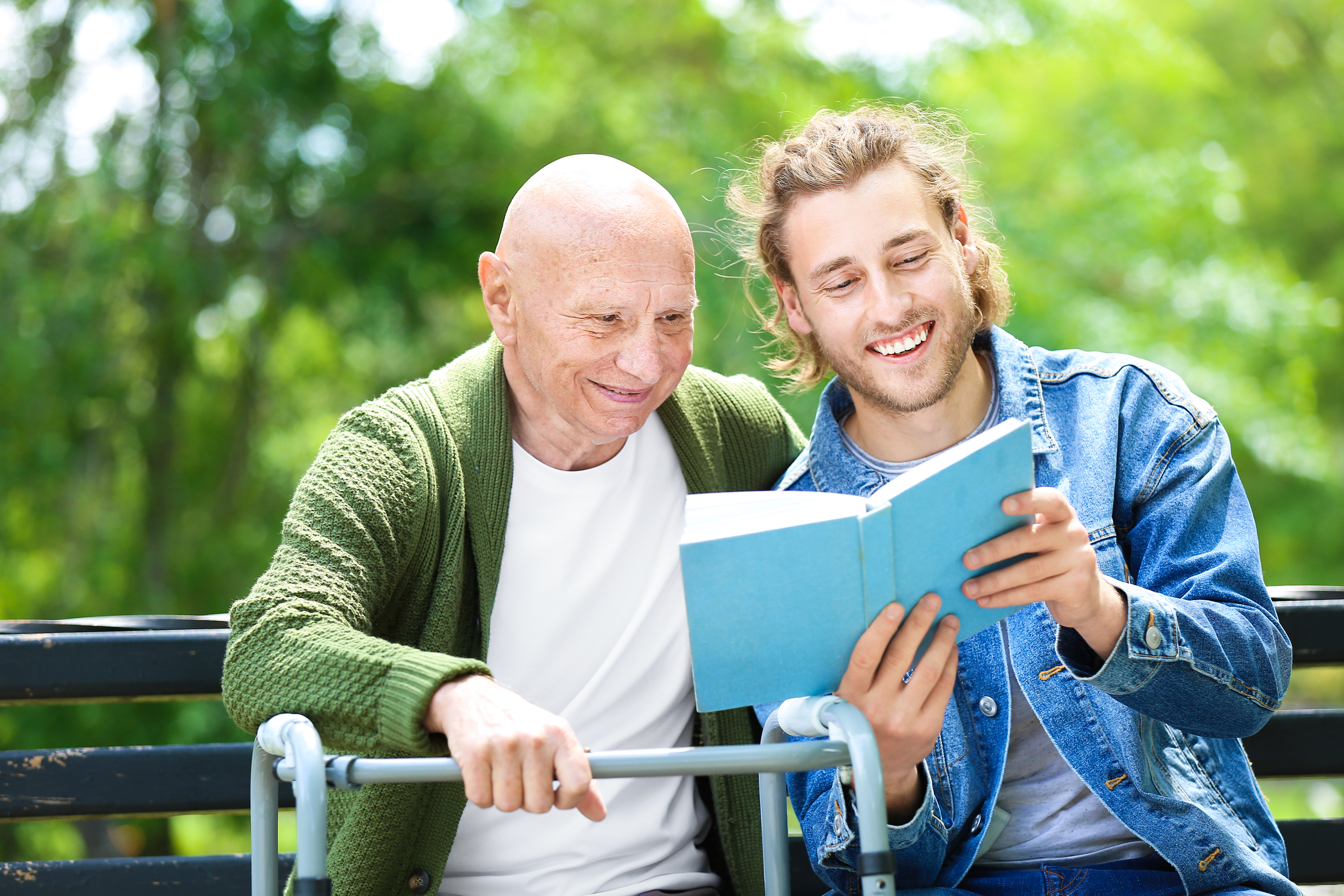 Adult man is holding a book in front of an older adult man, reading together on a bench. Older adult man is resting one hand on a walker.