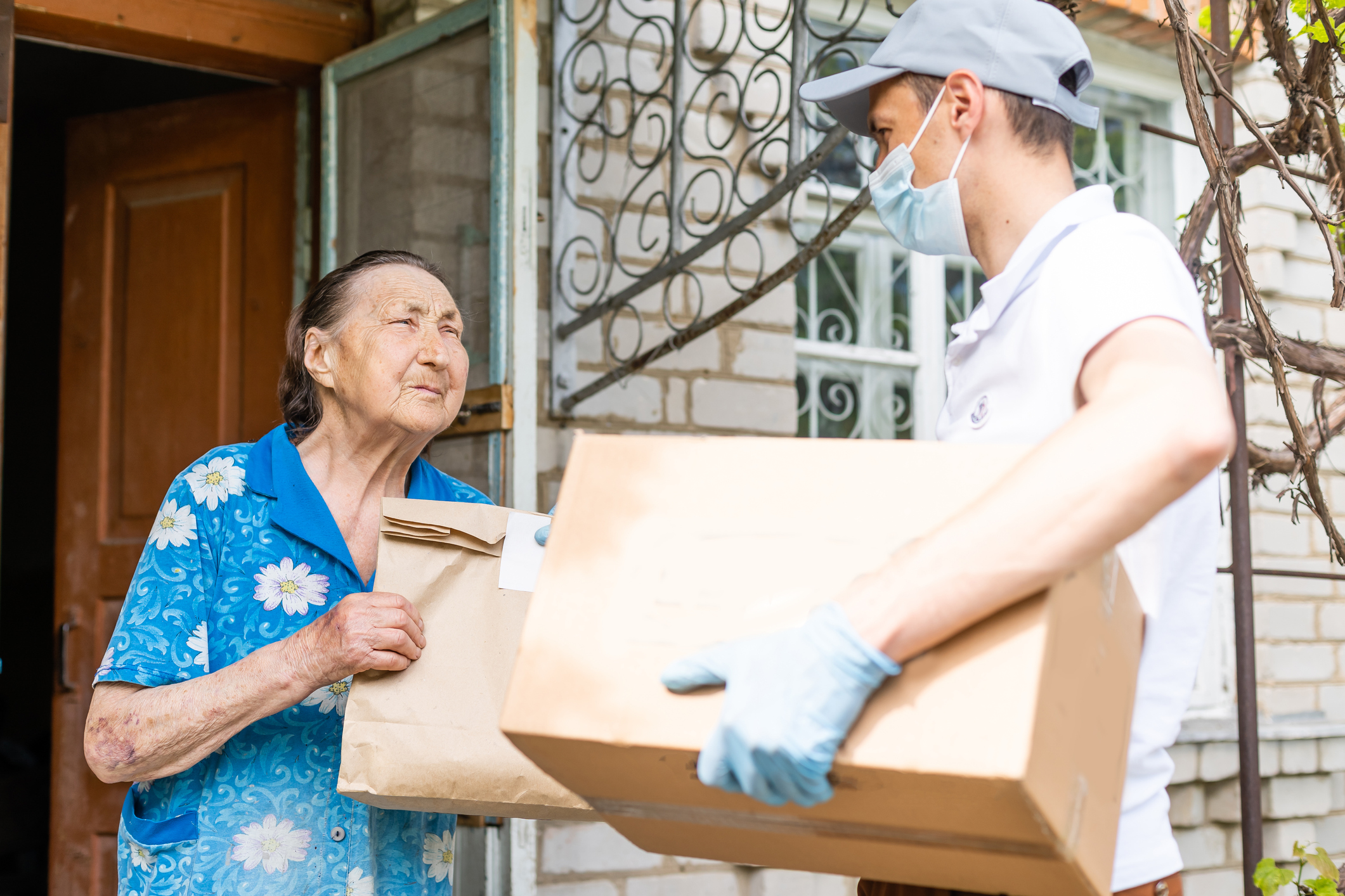 Young male volunteer in mask gives an elderly woman boxes with food near her house