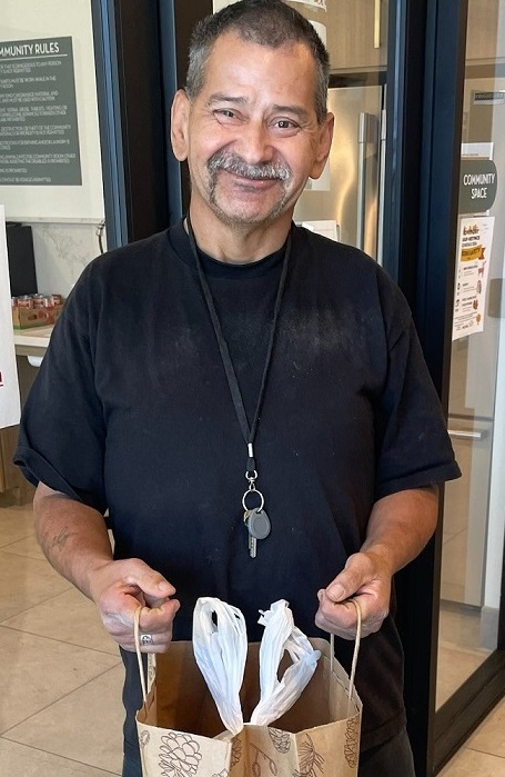 Jessie stands in front of a door, holding a bag of food with a smile on his face.