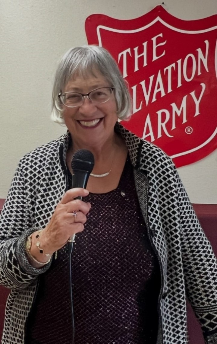 Marilyn Bernstone stands with a microphone in front of a Salvation Army sign, ready to speak.