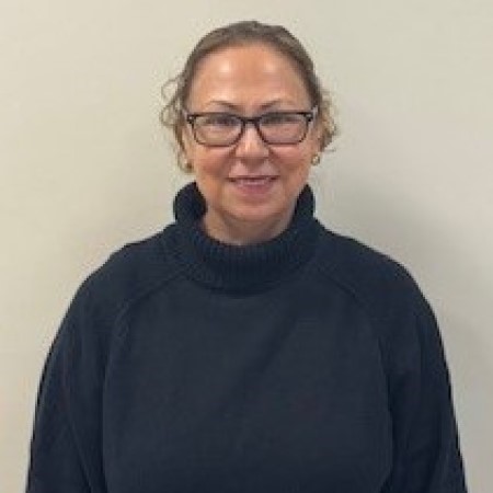 Gloria Villalpando wearing a black sweater stands in front of a plain wall, looking directly at the camera.