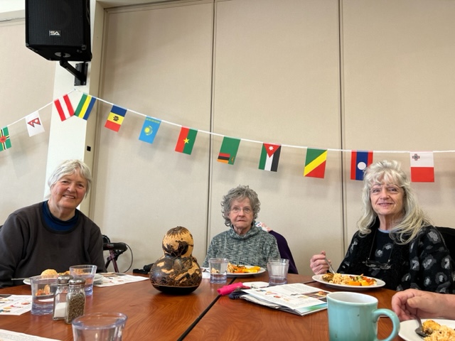 (Anne-Nancy-Jillian) Three women are eating a meal together, seated under a banner of international flags with a musical instrument on the table.