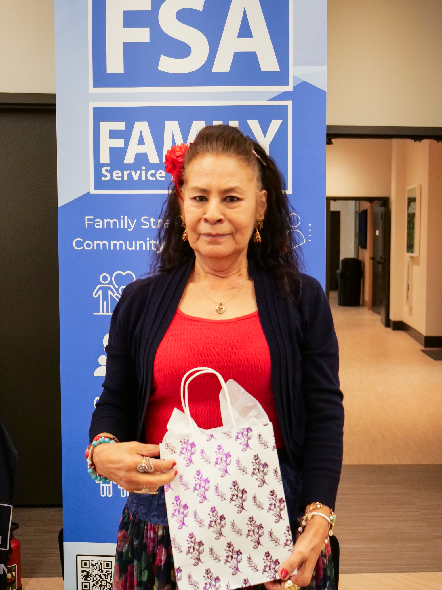 Older adult woman with dark hair, a red flower in her hair and red shirt with black cardigan holding gift bag in front of a sign for the Family Services Association