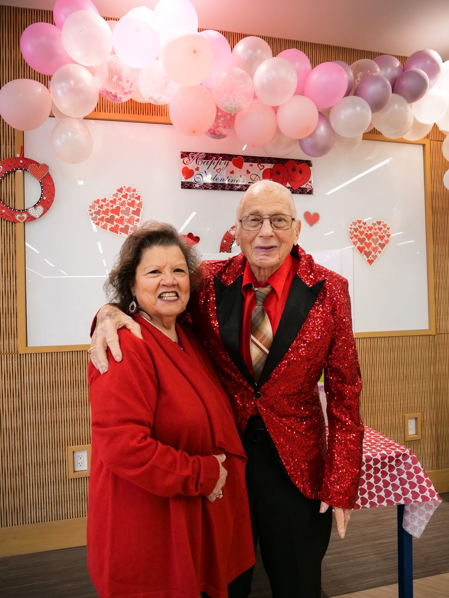 Older adult man in red sparkling blazer with his arm around an older adult woman wearing a red cardigan