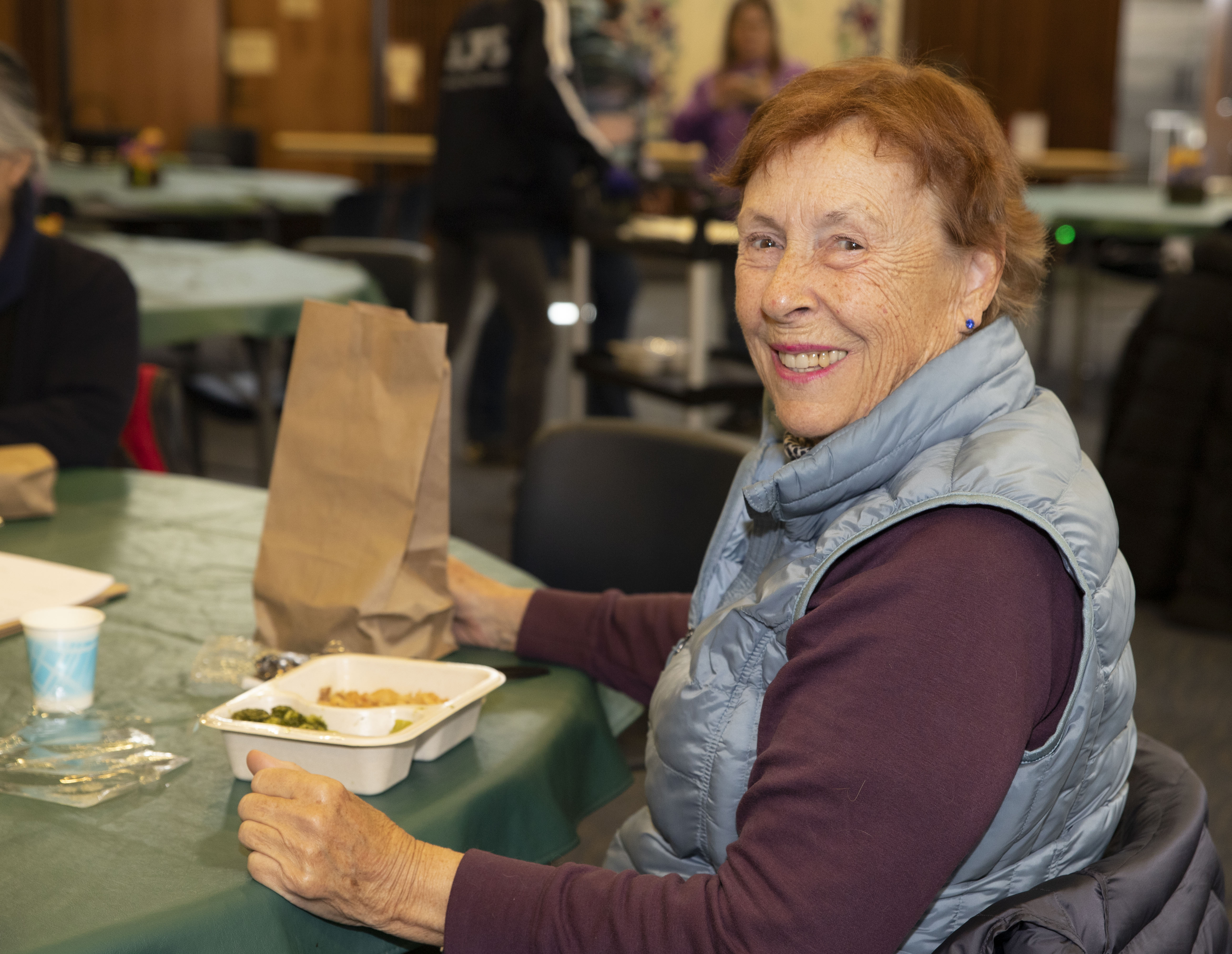 Woman with red hair in maroon long sleeve shirt and blue vest smiling with her meal