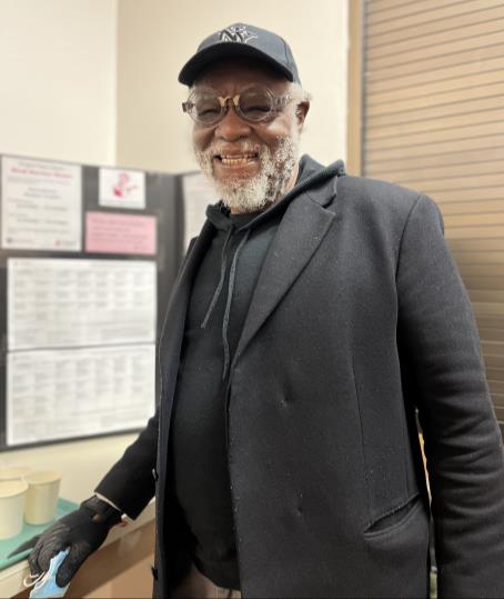 Smiling white-bearded man with clear frame glasses, black coat and black hoodie