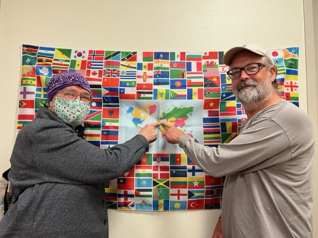 (Trina-Pierre) – A woman wearing a purple bandana, glasses, a mask and dark grey sweatshirt and man in glasses, a cap and tan long sleeve shirt point to poster of a world map with international flags